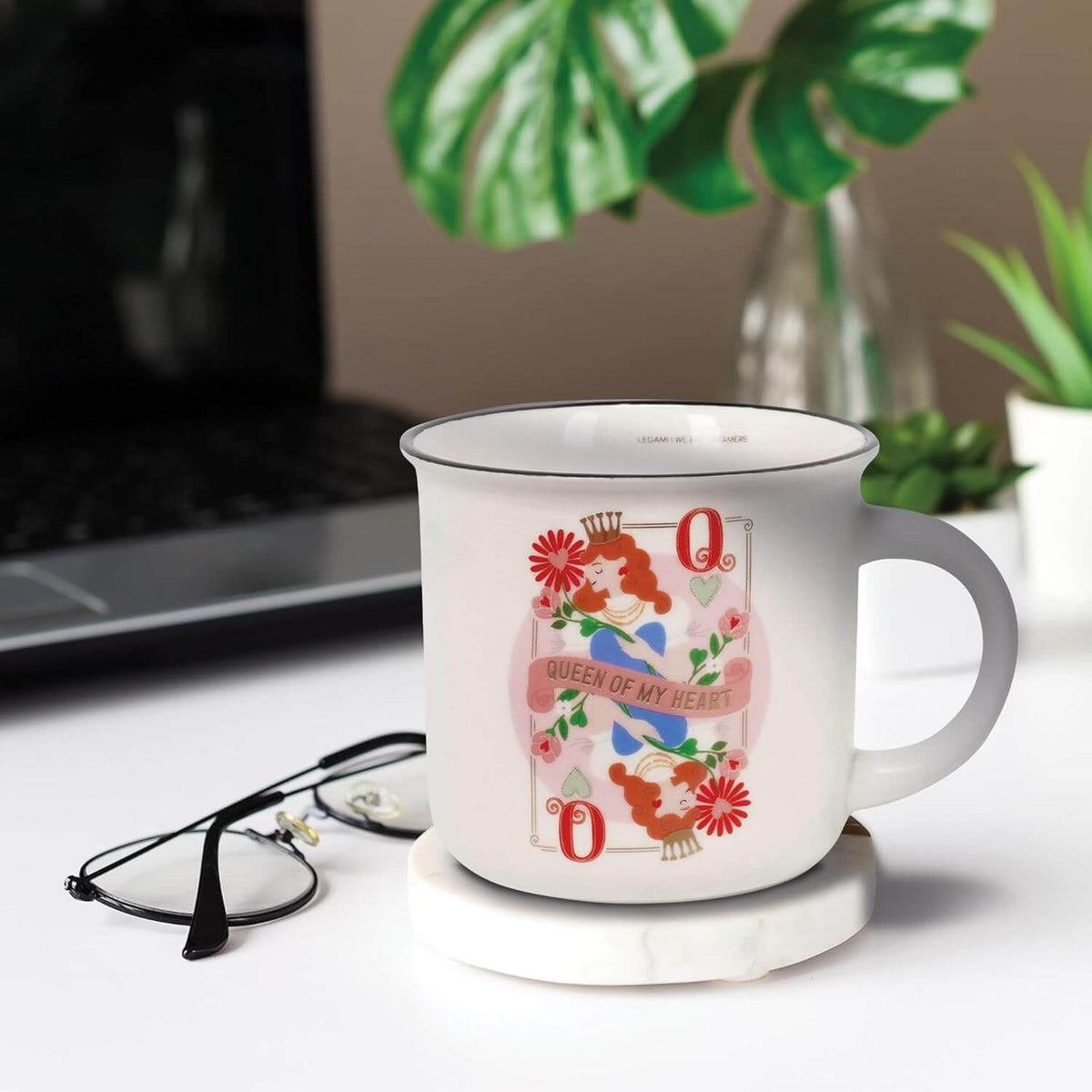 Mug da caffè e tè Legami Queen su un sottobicchiere in marmo accanto a occhiali e computer portatile